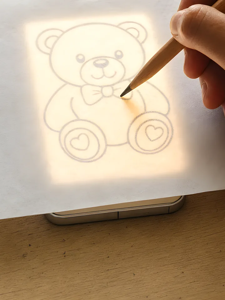 A teddy bear outline glowing through a sheet of paper placed on a phone screen